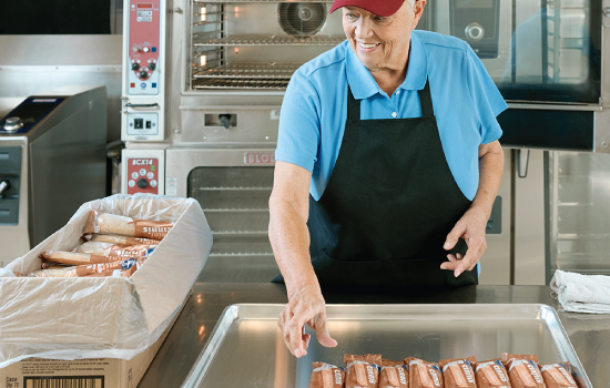 Two cafeteria workers in aprons and hairnets standing in a commercial kitchen, one holding a tray of food.